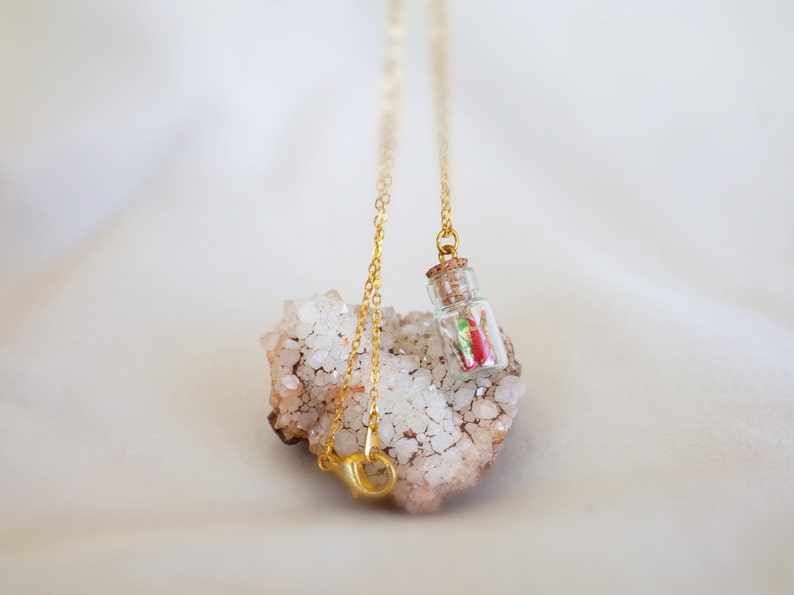 A necklace with a miniature glass bottle containing a tiny red and green origami crane, suspended on a golden chain, delicately resting against a cluster of sparkling white crystals.