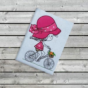 Könnte beinhalten: Gesticktes Design eines Mädchens auf einem Fahrrad, das einen rosa Hut mit weißen Tupfen und ein passendes Kleid trägt. Das Fahrrad hat einen Korb mit Blumen. Das Kunstwerk befindet sich auf hellblauem Hintergrund.