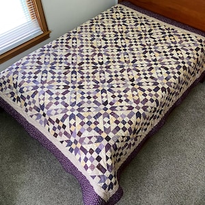 May include: A handmade quilt with a geometric pattern in shades of purple, cream, and brown covers a bed. The quilt features a repeating diamond and square design. The border is a solid purple with a dotted pattern.