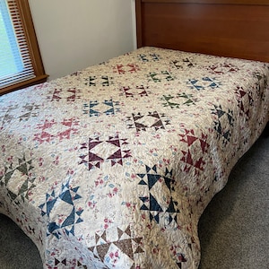 May include: A white quilt with a floral pattern and a patchwork design featuring blue, red, and brown squares. The quilt is on a bed with a wooden headboard.