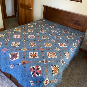 May include: A blue quilt with a patchwork design featuring various geometric shapes and colors, including red, pink, green, brown, and white. The quilt is laid out on a bed with a wooden headboard.
