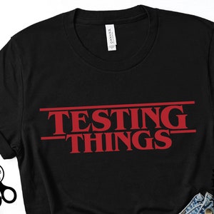 Op de afbeelding: Zwart T-shirt met rode tekst die "Testing Things" zegt in een stijl die doet denken aan de populaire serie "Stranger Things".