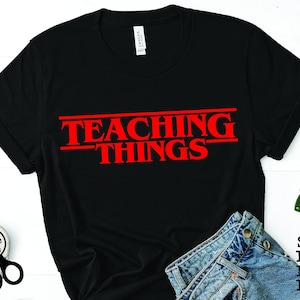 Pode incluir: Camiseta preta com texto vermelho que diz "TEACHING THINGS" em uma fonte estilizada que lembra a popular série de televisão Stranger Things.