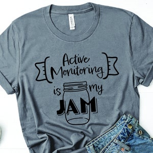 May include: A gray t-shirt with the text "Active Monitoring is my JAM" in black. The text is surrounded by a black arrow design. A black and white illustration of a mason jar is in the center of the text.