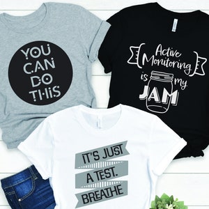 May include: Three t-shirts with different designs. The first t-shirt is gray with black text that says "You Can Do This". The second t-shirt is black with white text that says "Active Monitoring is my Jam" and a drawing of a jar. The third t-shirt is white with gray text that says "It's Just A Test. Breathe".