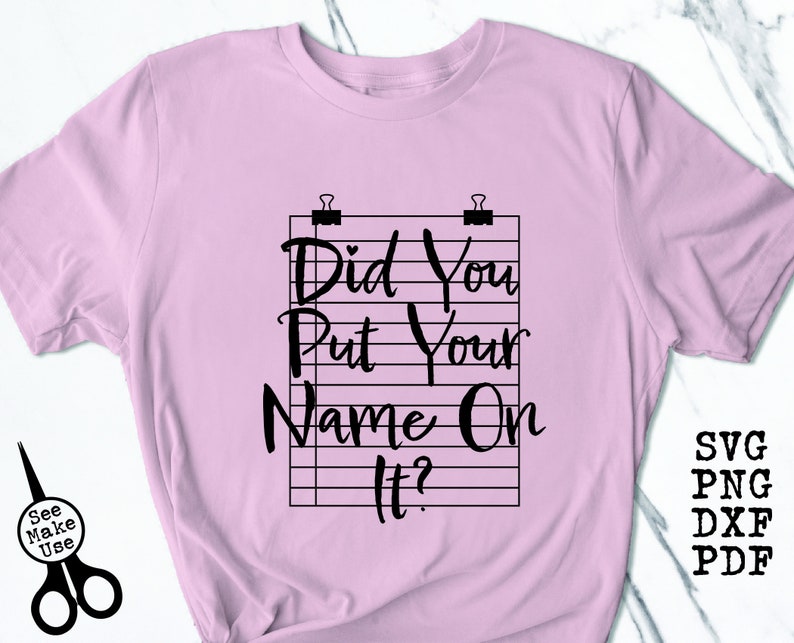 Pode incluir: Camiseta rosa com um gr&aacute;fico preto que diz "Did You Put Your Name On It?" O gr&aacute;fico &eacute; uma representa&ccedil;&atilde;o estilizada de uma folha de papel pautada com clipes de papel no topo.
