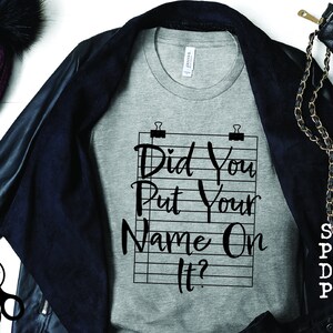 Pode incluir: Uma camiseta cinza com o texto "Did You Put Your Name On It?" impresso nela. O texto est&aacute; em uma fonte preta, escrita &agrave; m&atilde;o e &eacute; cercado por uma grade preta e branca.