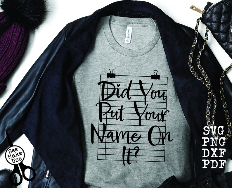 Pode incluir: Uma camiseta cinza com o texto "Did You Put Your Name On It?" impresso nela. O texto est&aacute; em uma fonte preta, escrita &agrave; m&atilde;o e &eacute; cercado por uma grade preta e branca.