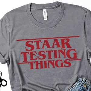 Puede incluir: Camiseta gris con el texto "STAAR TESTING THINGS" en letras rojas, estilizado para parecerse al logotipo de Stranger Things.