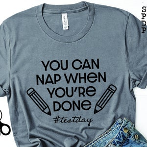 Op de afbeelding: Een grijs T-shirt met zwarte tekst die "You can nap when you're done #testday" luidt. Twee zwarte potloodschetsen bevinden zich aan beide kanten van de tekst.