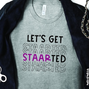 Op de afbeelding: Grijs T-shirt met de tekst "Let's Get Staarted" in zwarte en roze letters. Het woord "Staarted" wordt meerdere keren herhaald met de letters "Staar" in roze.