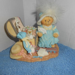 Puede incluir: Una figurita de un oso de peluche vestido con un vestido azul y un sombrero blanco con una pluma. El oso sostiene un abanico y una cadena de perlas. El oso está de pie junto a una pequeña maleta abierta llena de varios artículos, incluyendo un sombrero, un paraguas y un pequeño oso de peluche.