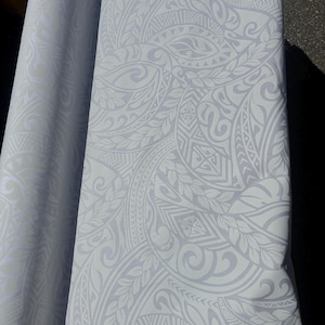 May include: A roll of fabric with a repeating, intricate pattern in shades of white and light gray. The design features swirling shapes, leaves, and geometric elements, creating a textured appearance. The fabric is folded over, revealing the pattern's continuity.
