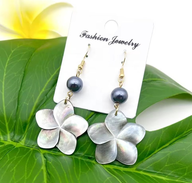 May include: A pair of dangle earrings featuring a black pearl and a white shell flower. The earrings are on a white card with the text "Fashion Jewelry".