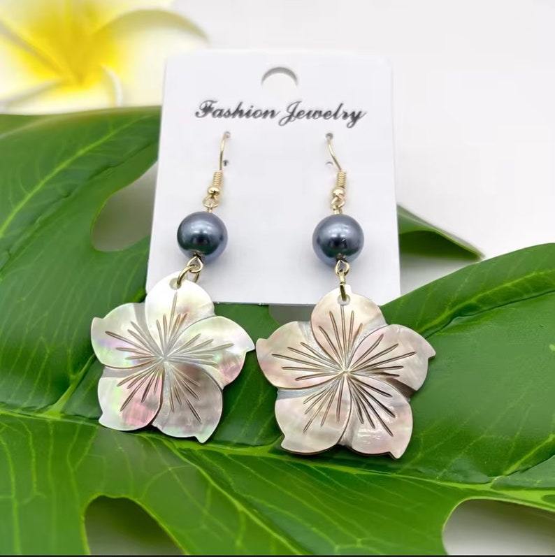 May include: A pair of gold dangle earrings featuring a flower-shaped shell pendant with a black pearl accent. The earrings are on a white card with the text "Fashion Jewelry".