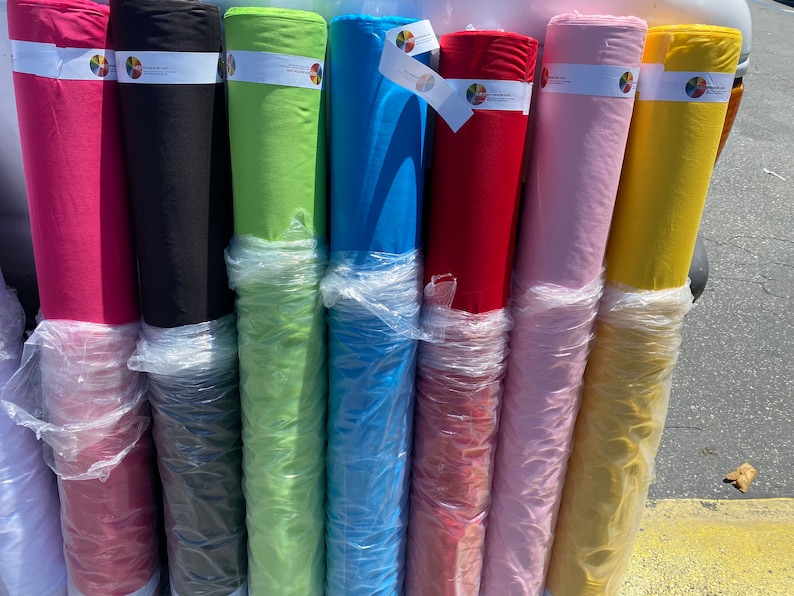 May include: Rolls of fabric in various colors, including pink, black, green, blue, red, light pink, and yellow. The fabric is wrapped in clear plastic and labeled with a colorful circle logo.