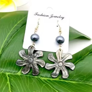May include: A pair of earrings featuring a black pearl and a silver flower-shaped charm. The earrings are hanging from a gold hook.