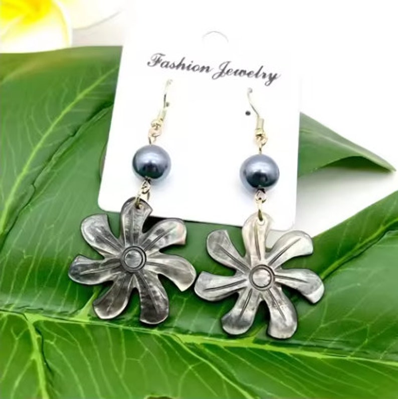 May include: A pair of earrings featuring a black pearl and a silver flower-shaped charm. The earrings are hanging from a gold hook.