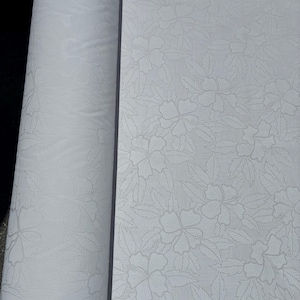 May include: A roll of white fabric featuring a tonal jacquard pattern of hibiscus flowers and leaves. The design includes subtle vertical stripes integrated into the background, creating a monochromatic floral motif across the entire surface.