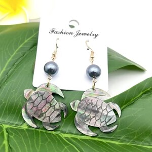 May include: A pair of dangle earrings featuring a black pearl and a turtle shell design. The turtle shell is made of mother of pearl and has an iridescent finish. The earrings are gold-toned and have a hook closure.