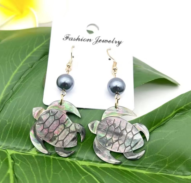 May include: A pair of dangle earrings featuring a black pearl and a turtle shell design. The turtle shell is made of mother of pearl and has an iridescent finish. The earrings are gold-toned and have a hook closure.