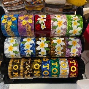 May include: A stack of colorful bangle bracelets with floral and geometric designs. The bracelets are made of plastic and have a glossy finish. The top row of bracelets features white plumeria flowers on a variety of backgrounds. The bottom row of bracelets features the word "TONG" in gold lettering on a variety of backgrounds.