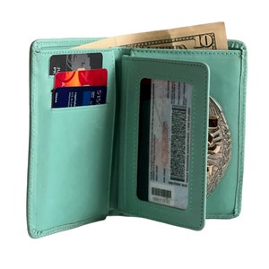 May include: Open, light green leather wallet with multiple card slots, a clear ID window, and a compartment holding a ten-dollar bill. The wallet also features a silver badge. The wallet is open, displaying its contents.
