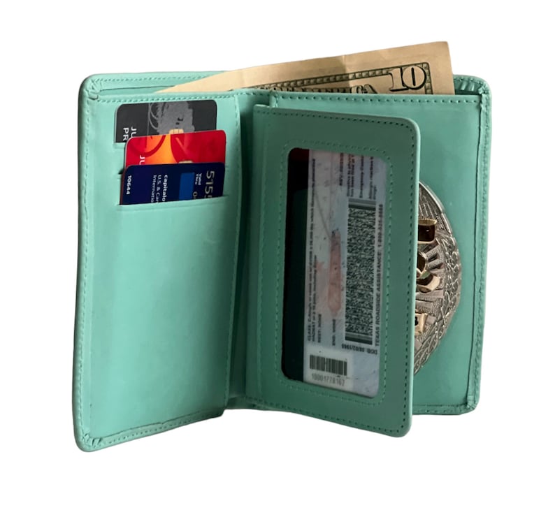 May include: Open, light green leather wallet with multiple card slots, a clear ID window, and a compartment holding a ten-dollar bill. The wallet also features a silver badge. The wallet is open, displaying its contents.