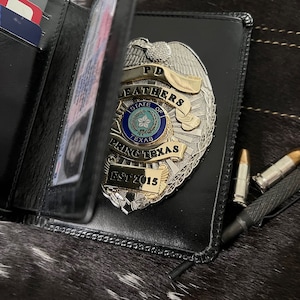 May include: Black leather wallet with a silver and gold police badge. The badge reads "PD Leathers State of Texas Spring Texas EST 2015." Two bullets and a black pen are next to the wallet. The wallet is open, revealing cards.