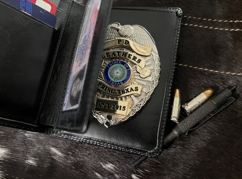 May include: Black leather wallet with a silver and gold police badge. The badge reads "PD Leathers State of Texas Spring Texas EST 2015." Two bullets and a black pen are next to the wallet. The wallet is open, revealing cards.