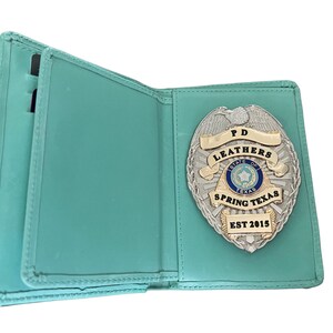 May include: Open, teal-colored leather badge holder with a silver and gold police badge. The badge features the text "PD LEATHERS STATE OF TEXAS SPRING TEXAS EST 2015". The holder has slots for cards.