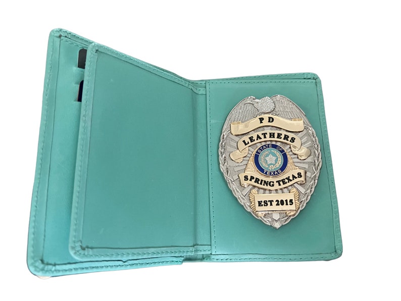 May include: Open, teal-colored leather badge holder with a silver and gold police badge. The badge features the text "PD LEATHERS STATE OF TEXAS SPRING TEXAS EST 2015". The holder has slots for cards.