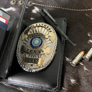 May include: A black leather wallet with credit cards, a silver and gold police badge with the text "SPRING TEXAS 2015", a black pen, and two bullets. The items are on a cowhide surface.