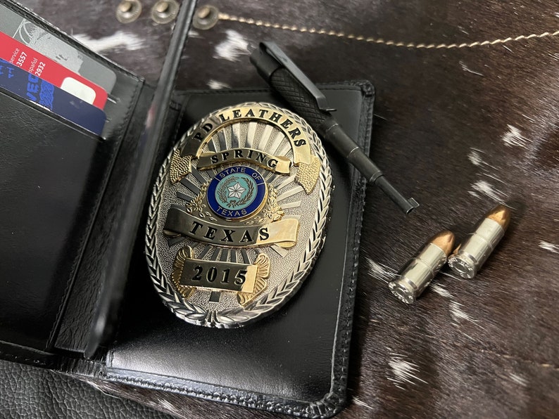 May include: A black leather wallet with credit cards, a silver and gold police badge with the text "SPRING TEXAS 2015", a black pen, and two bullets. The items are on a cowhide surface.