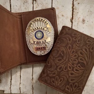 Può includere: Un portafoglio in pelle marrone con un motivo floreale in rilievo accanto a un portafoglio in pelle marrone aperto con un distintivo PD LEATHERS SPRING TEXAS 2015. Il distintivo è argentato e dorato con accenti blu e bianchi.