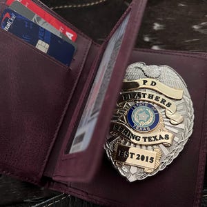 May include: A burgundy leather wallet with a silver and gold police badge. The badge reads "PD Leathers State of Texas Spring Texas EST 2015." Credit cards are visible inside the wallet.