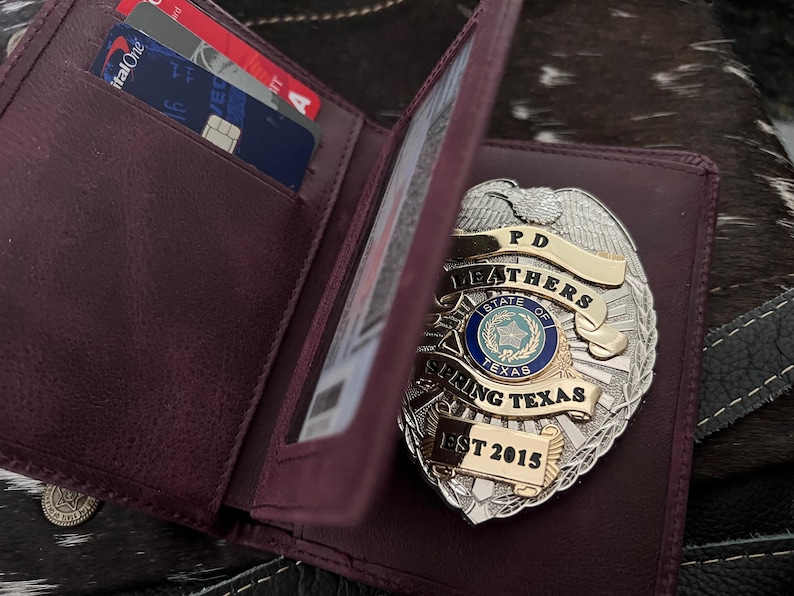 May include: A burgundy leather wallet with a silver and gold police badge. The badge reads "PD Leathers State of Texas Spring Texas EST 2015." Credit cards are visible inside the wallet.