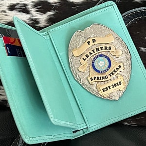 May include: A teal leather badge holder with a silver and gold police badge. The badge reads "PD LEATHERS STATE OF TEXAS SPRING TEXAS EST 2015". Credit cards are visible inside the holder.