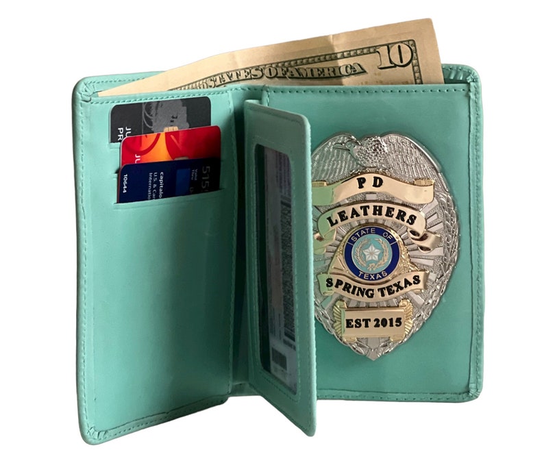 May include: Open, light teal leather wallet with a silver badge that reads "PD LEATHERS SPRING TEXAS EST 2015." The wallet holds credit cards and a ten-dollar bill.