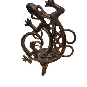 May include: A bronze-coloured metal lizard-shaped decorative object. The lizard has a detailed design with a textured body and outstretched limbs. The tail curls around a circular element with three loops. Likely a decorative piece or wall hanging.
