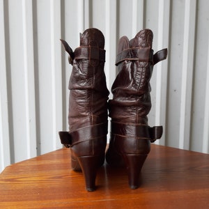 Pu&ograve; includere: Un paio di stivali in pelle marrone con tacco medio. Gli stivali hanno un design stropicciato e sono assicurati con cinturini con fibbia. Gli stivali sono esposti su una superficie di legno.