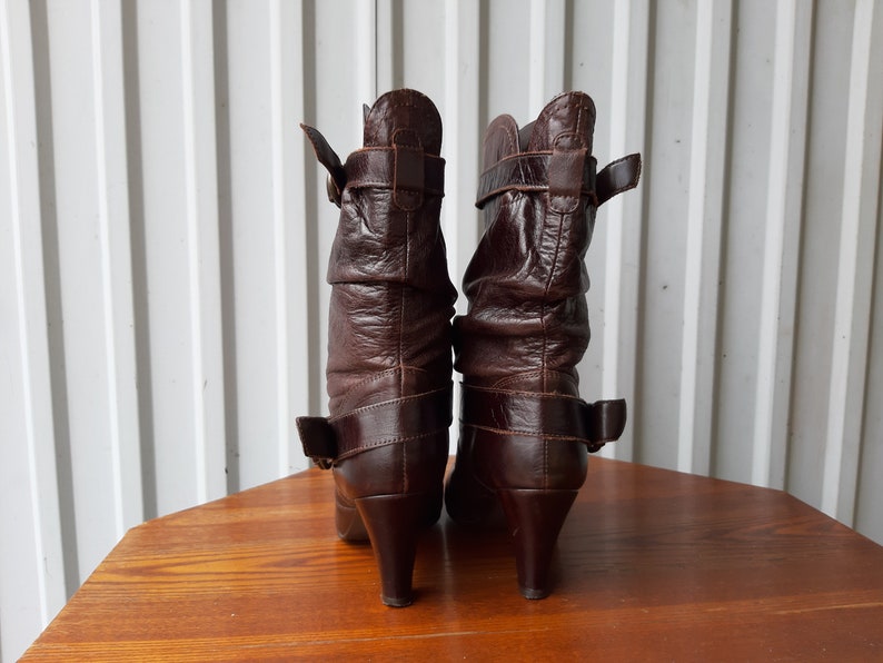 Pu&ograve; includere: Un paio di stivali in pelle marrone con tacco medio. Gli stivali hanno un design stropicciato e sono assicurati con cinturini con fibbia. Gli stivali sono esposti su una superficie di legno.
