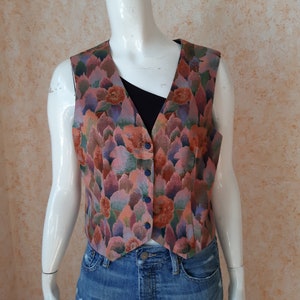 Puede incluir: Un chaleco con estampado floral con cuello en V y cierre de botones. El chaleco presenta un patrón de flores y hojas rosas, naranjas, verdes y moradas. Se muestra en un maniquí, combinado con una camiseta negra y vaqueros azules.