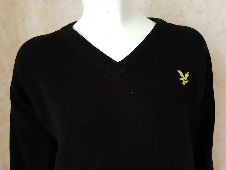 May include: Black V-neck sweater with long sleeves. The sweater features a small yellow embroidered eagle logo on the chest. The knitwear is displayed on a white mannequin.