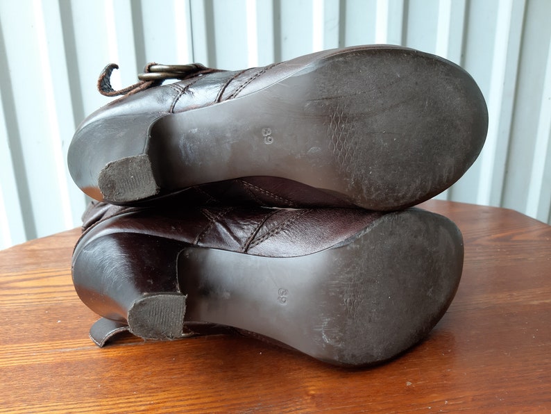 Pu&ograve; includere: Un paio di stivaletti alla caviglia in pelle marrone con tacco. Gli stivaletti hanno un dettaglio con fibbia e sono taglia 39. Le suole mostrano segni di usura. Gli stivaletti sono su una superficie di legno.