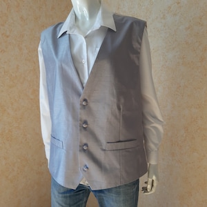 May include: A light gray vest with a subtle sheen, featuring a button-up front and two front pockets. The vest is worn over a white collared shirt and blue jeans. The vest is a formal garment.