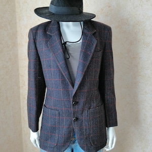May include: A navy blue blazer with a red plaid pattern, two front pockets, and black buttons. A gray fedora hat sits above the blazer on a mannequin. The blazer is part of a vintage clothing collection.