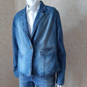 May include: A light blue denim blazer with a single button closure and two front pockets. The blazer is worn over a white collared shirt and paired with matching denim jeans. The jacket has a classic design.