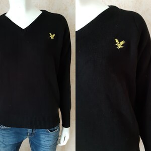 May include: Black V-neck sweater with long sleeves. A gold-colored bird emblem is embroidered on the left chest. The sweater is made of a knit fabric and is displayed on a mannequin.