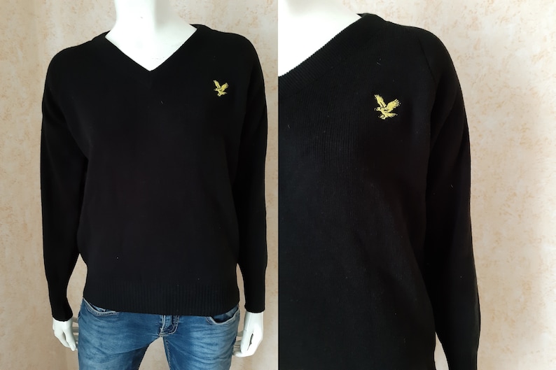 May include: Black V-neck sweater with long sleeves. A gold-colored bird emblem is embroidered on the left chest. The sweater is made of a knit fabric and is displayed on a mannequin.